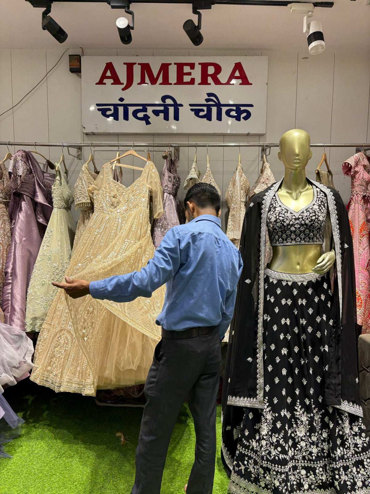 An Ajmera staff member showing a cream lehenga beside a mannequin in a black lehenga, under the Ajmera sign